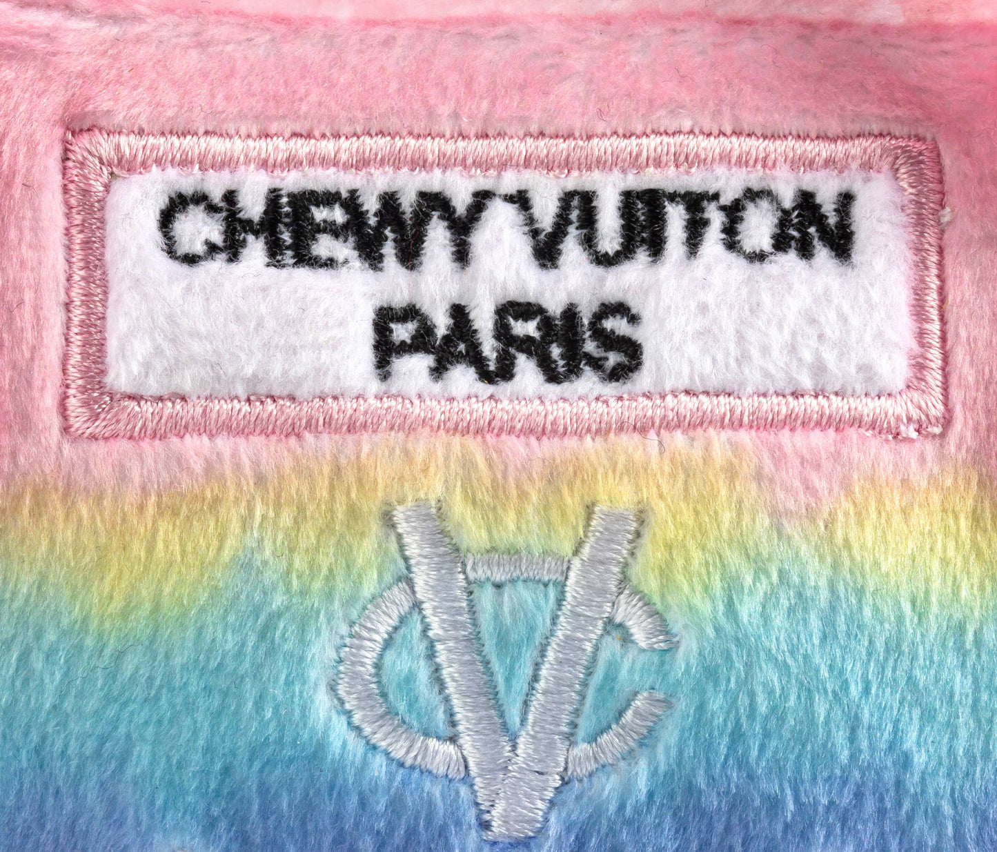 Plush dog toy shaped like a designer handbag with pink handles and a pastel rainbow gradient featuring embroidered icons and a “Chewy Vuiton Paris” label. Soft luxury squeaky chew toy for pets.