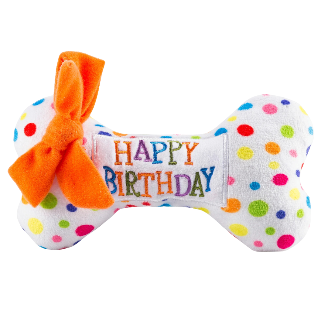 White plush dog bone toy with colorful polka dots, an orange bow, and embroidered “Happy Birthday” text. Soft squeaky celebration chew toy for pets.