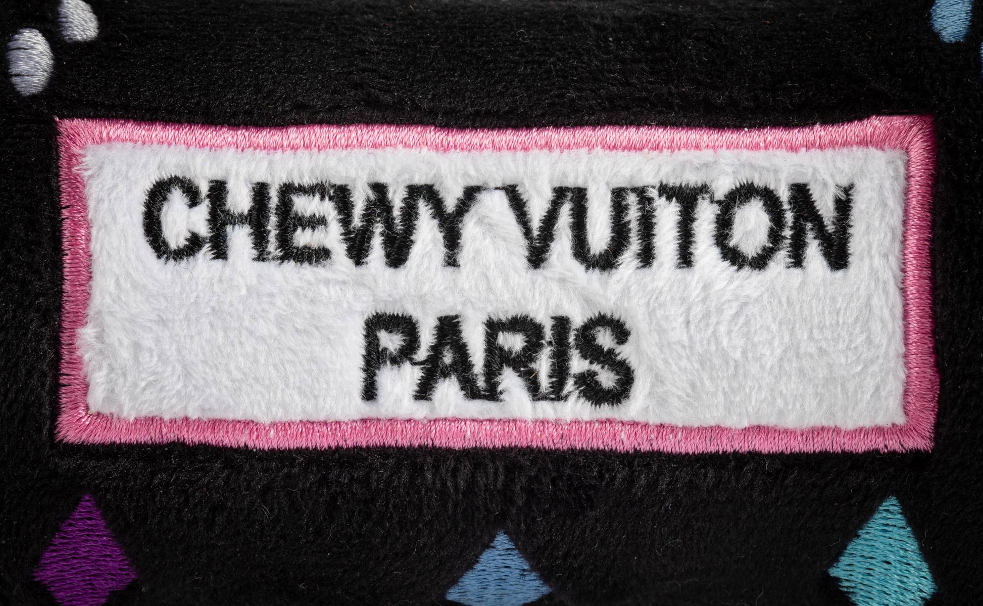 Black plush dog bone toy with colorful embroidered designer-style icons and a “Chewy Vuiton Paris” label. Soft luxury squeaky chew toy for pets.