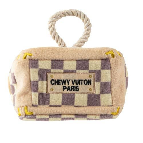 Plush interactive dog toy shaped like a designer-style trunk with a beige and lavender check pattern, rope handle, and “Chewy Vuiton Paris” label, including four matching plush balls to hide and retrieve.