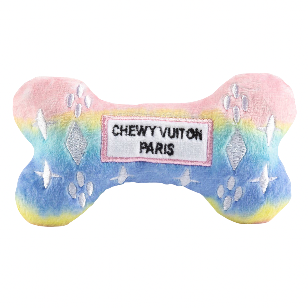 Pastel rainbow plush dog bone toy in pink, blue, and yellow gradient with white embroidered icons and a “Chewy Vuiton Paris” label. Soft luxury squeaky chew toy for pets.