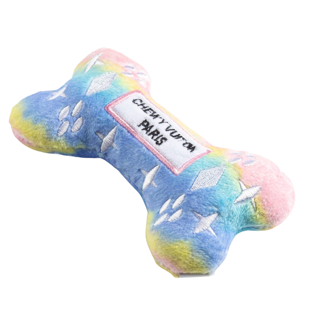 Pastel rainbow plush dog bone toy in pink, blue, and yellow gradient with white embroidered icons and a “Chewy Vuiton Paris” label. Soft luxury squeaky chew toy for pets.
