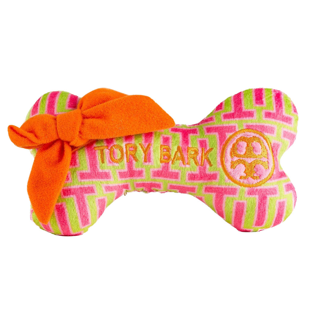 Plush dog bone toy with pink and green geometric pattern, orange bow, and embroidered “Tory Bark” label with matching logo. Soft luxury squeaky chew toy for pets.
